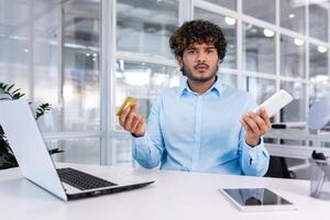 Debt, bankruptcy, foreclosure, fraud. A young Latin American businessman is sitting in the office at the table, holding a phone and a credit card in his hands. He looks worriedly at the camera. photo