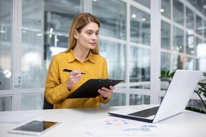 An attractive blonde woman works in a modern office, sits at a desk in front of a notebook, does paperwork, looks intently at the computer screen, jots down information. photo