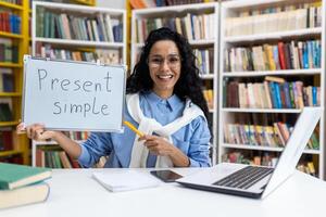 Cheerful female teacher holding a board with 'Present Simple' written on it, demonstrating English grammar in a library setting surrounded by books. photo