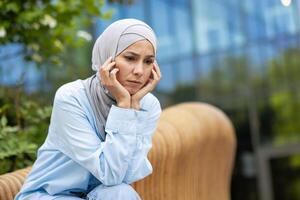 solitario joven mujer en hijab sentado pensativamente en un banco en un pacífico urbano parque, encarnando serenidad y introspección. foto