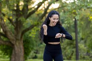 alegre Hispano mujer comprobación su aptitud rastreador después un vigoroso correr en un sereno parque configuración, expresando felicidad y satisfacción con su progreso. foto