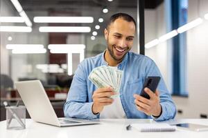 sonriente Hispano joven hombre sentado en un formal oficina a un mesa, participación efectivo dinero billetes en su manos, utilizando un móvil teléfono. foto