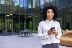 retrato de un joven africano americano mujer de negocios en pie fuera de un oficina centro, participación un móvil teléfono y sonriente a el cámara. foto