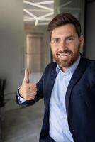 Vertical close-up photo of a young smiling and successful man working in the office and talking on a call on the phone, showing the super success sign to the camera.