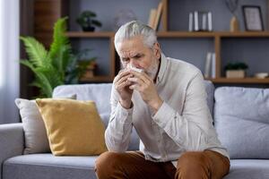 un mayor hombre experimentando síntomas de un frío o gripe, sentado en un sofá y utilizando un pañuelo de papel en un moderno, acogedor vivo habitación. foto