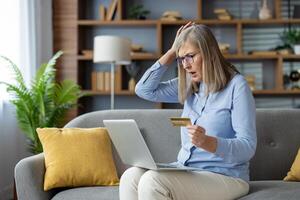 A mature woman looking surprised and anxious while holding a credit card and using a laptop on her sofa at home. photo