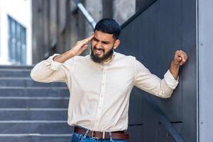 A sick Indian young man stands outside the building on the steps and holds his head with his hand, twisted in pain, feeling great pressure and tension photo