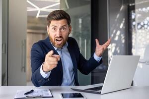 enojado joven hombre empresario sentado en el oficina a el mesa y señalando su dedo a el cámara, consiguiendo usado a el problema y error, gritos disgustado. foto