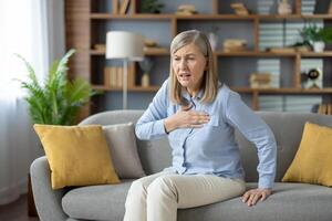 Elderly female grabbing chest with palm while sitting on pillowed couch with shocked expression. Ill woman feeling sharp cardiac pain and risk of infarction while sitting alone in modern flat. photo