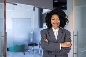 Portrait of young African American businesswoman, woman with crossed arms smiling and looking at camera, female worker at workplace inside office, boss in business suit smiling with satisfaction. photo