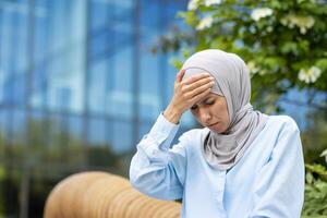 Sick woman in grey headscarf placing hand on forehead while sitting with closed eyes in bad condition. Stressed lady suffering from headache and sunstroke during unexpected heat in springtime. photo