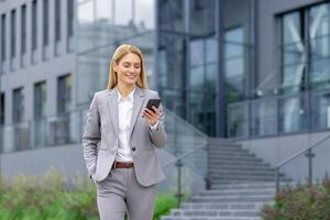 Successful woman boss walks outside office building outdoors, in business suit, businesswoman uses app on phone, smiles contentedly reads internet pages, browses news, writes text message, call. photo