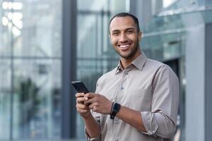 Portrait of African American businessman, man holding phone smiling and looking at camera, office worker in shirt outside modern office building on lunch break photo