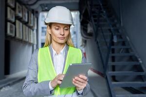 Serious confident thinking female engineer with tablet computer inspecting factory wearing hard hat and reflective vest. photo