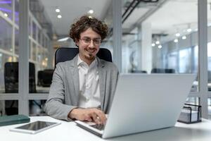 Young, professional male office worker with curly hair typing on a laptop, exhibiting efficiency and a contented demeanor amidst the corporate hustle. photo