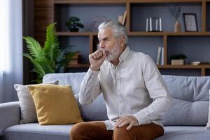 Mature man feeling unwell, coughing while sitting alone on a sofa in a well-decorated, contemporary living room. Concept of aging and health issues. photo