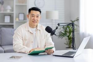 un enfocado hombre en un elegante, brillante hogar oficina preparar absorto en un libro con un podcasting micrófono montado en el escritorio. foto