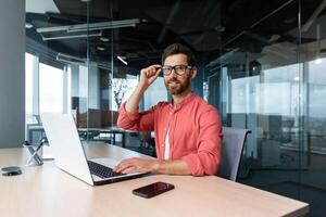 Portrait of mature businessman freelancer startup, bearded man smiling and looking at camera, business owner working inside modern office building wearing red shirt and glasses. photo