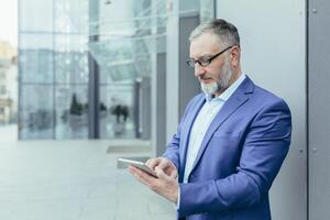 Senior handsome businessman man in glasses and a suit stands on the street near the office center and waits for customers, employees, holds the phone in his hands, dials a number, messages. photo