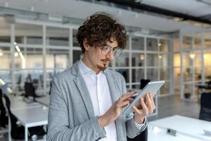 pelo rizado empresario en un traje chaqueta absorto en su tableta trabajo a un brillante corporativo oficina. foto