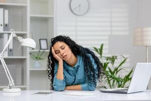 An overworked businesswoman taking a nap at her workspace, with a laptop and phone, in a modern office setting. photo