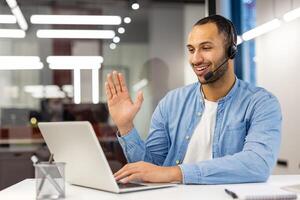 un joven Hispano hombre en un traje es sentado en el oficina a un ordenador portátil en un auriculares y hablando en un llamar, sonriente y ondulación a el cámara. foto