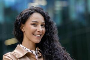 de cerca retrato de joven hermosa Hispano mujer de negocios sonriente y mirando a cámara con Rizado pelo fuera de oficina edificio, financiero contento con exitoso logro resultados. foto