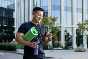 A young Asian sportsman man is standing on a city street in headphones, holding a phone, a yoga mat, and a bottle of water. photo