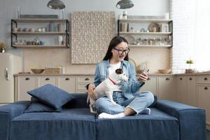 joven hermosa asiático mujer con mascota Jack Russell terrier hablando en llamada con amigos, mujer a hogar en cocina utilizando teléfono inteligente para remoto comunicación. foto