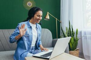 Happy young hispanic woman in headphones talking looking at laptop, talking via conference, teacher teaching via webcam, online learning, greeting, waving. photo