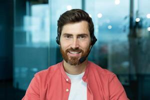 Close-up portrait of mature man with beard and call headset inside office, tech support worker smiling and looking at camera in red shirt, online customer service and support. photo