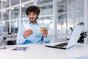 en línea compras, pago y comprobación de cuentas un joven Hispano hombre es sentado en el oficina a un escritorio con un computadora portátil, participación un teléfono y utilizando un crédito tarjeta. foto