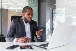 Angry nervous businessman talking remotely on call using laptop, mature experienced boss shouting and arguing sitting at workplace inside office. photo