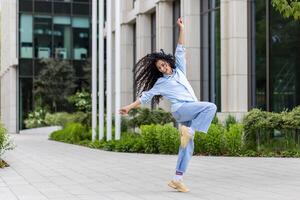 A youthful and vibrant woman is captured in mid-dance, radiating happiness and freedom against the modern architecture of an urban environment. Her dynamic movement and joyful expression exemplify carefree urban living and the celebration of life. photo