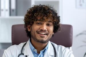 de cerca retrato de médico dentro clínica a lugar de trabajo, hombre en blanco médico Saco sonriente mirando a cámara, satisfecho indio hombre con Rizado cabello. foto