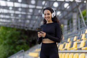 un joven mujer en atlético vestir con auriculares participación un teléfono inteligente mientras en pie en un estadio. concepto de aptitud y moderno estilo de vida. foto