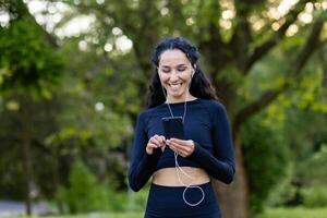 un alegre Hispano mujer escucha a música en su teléfono inteligente mientras corriendo en un lozano verde parque. ella usa un negro Deportes atuendo y exuda felicidad y salud. foto