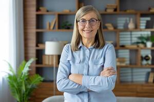 Confident female pensioner in pastel blouse standing with arms crossed on chest and smiling at camera. Mature lady in spectacles posing at stylish apartment interior and enjoying holiday indoors. photo