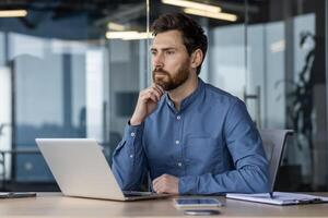 A serious young man businessman sits in the office at a desk with a laptop, holds his hand on his chin and looks thoughtfully to the side. photo