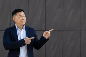 asiático joven masculino empresario en pie cerca un oficina edificio en un gris pared antecedentes en un traje y sonriente señalando con su dedos en un gris muro, blanco antecedentes Bosquejo. foto