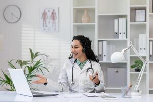 un profesional hembra médico con un estetoscopio comprometido en un interactivo en línea consulta, sonriente calurosamente en un bien equipado médico oficina. foto