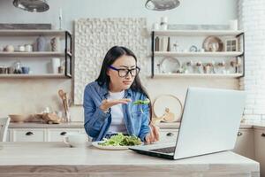 insatisfecho asiático mujer con en línea dietas, mujer sentado en cocina a hogar comiendo ensalada para peso pérdida, utilizando ordenador portátil a ver recomendaciones y aprender acerca de dieta y sano comiendo. foto