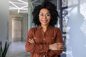 retrato de exitoso oficina obrero, africano americano mujer con Rizado pelo sonriente y mirando a cámara, mujer de negocios con cruzado brazos a lugar de trabajo contento con logro resultados dentro oficina. foto