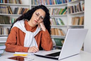 aburrido caucásico hembra sentado a escritorio con moderno ordenador portátil y trabajando con papel materiales con libros en frente de a ellos. trastornado mujer con lentes sensación cansado desde monótono trabajo proceso. foto