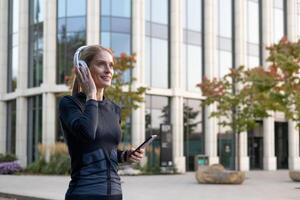 Joyful female in sporty outfit using smartphone and headphones outside modern buildings, radiating positivity and active lifestyle. photo