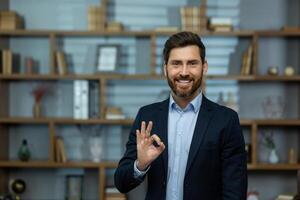 Professional mature male business owner showing positive approval with an okay hand sign in a well-organized office setting. photo