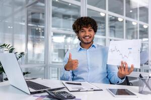 retrato de joven exitoso financiero papeleo dentro oficina, hombre sonriente y mirando a cámara muestra grafico con positivo dinámica de económico ganancias, empresario a trabajo con ordenador portátil. foto