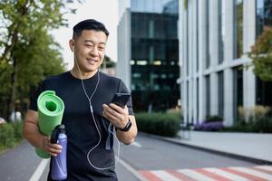 Young Asian male athlete in headphones goes to training on the city street. He holds a mat, a bottle of water in his hand and uses the phone with a smile. photo