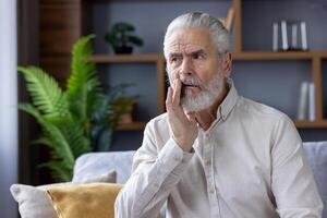 A concerned elderly man in a casual shirt sits on a sofa, touching his cheek due to a toothache, with a pained expression in a home setting. photo