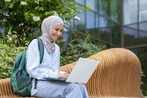 A cheerful Muslim woman wearing a hijab sits on a bench with a laptop, surrounded by greenery in a tranquil outdoor setting. photo